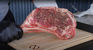 Hand in black glove seasoning a Tomahawk steak on a wooden Z branded cutting board, highlighting the rich marbling and generous coating of spices.