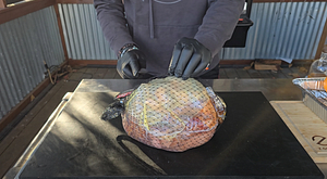 Chef wearing black gloves handling a packaged spiral ham on a cutting board, preparing it for seasoning and grilling.