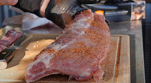 Preparing baby back ribs by sprinkling seasoning on a wooden cutting board, with a bag of Z Grills Cherry Wood Pellets in the background.