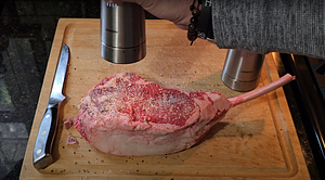 Seasoning a raw Tomahawk steak with salt and pepper on a wooden cutting board, preparing it for smoking and searing.