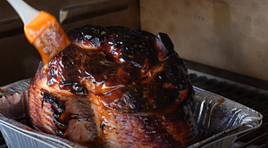 Glazing a smoked ham with a glossy maple bourbon sauce using a silicone brush, capturing the sauce dripping over the caramelized exterior.