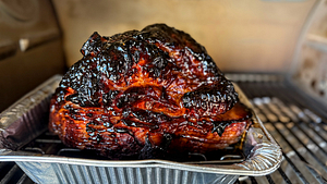 Close-up of a fully glazed and smoked ham resting in a foil tray on the grill, showcasing its glossy, caramelized crust.