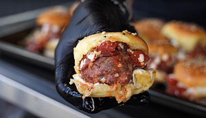 Close-up of a freshly prepared meatball slider with tomato sauce and melted cheese, held in a gloved hand.