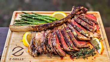 Sliced, perfectly cooked tomahawk steak served on a wooden cutting board with Z Grills logo, accompanied by fresh green asparagus and lemon slices, seasoned with herbs.