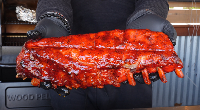 A person in black gloves holds a rack of glistening, freshly smoked ribs covered in a vibrant red BBQ sauce.